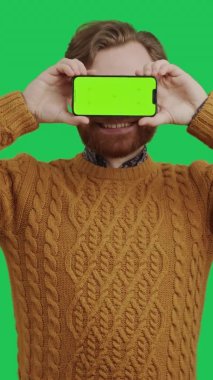 dikey çekim sakallı beyaz adam yeşil arka planda yeşil ekran akıllı telefonuyla gözlerini kapatıyor.