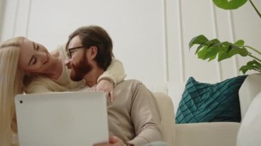 Kafkasyalı mutlu çift oturma odasında oturuyor ve gümüş bir dizüstü bilgisayar kullanarak internette geziniyorlar.
