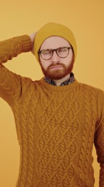 Hipster görünümlü beyaz adam ne yapacağını düşündü ve tek eliyle kafasını kaşıdı. Dikey stüdyo sarı arka plan.