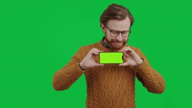 Orta yaşlı beyaz girişimci elinde yeşil ekranlı akıllı telefon ve yeşil arka planda gülümseyen orta büyüklükte bir stüdyo görüntüsü tutuyor.