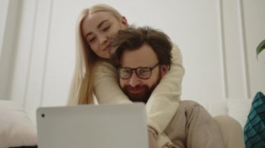 Sevecen beyaz bir çift oturma odasında oturuyor ve gümüş bir dizüstü bilgisayar kullanarak internette geziniyorlar.