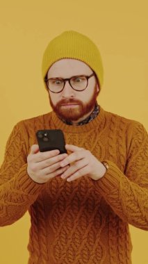 Kafkas sakallı bir adam akıllı telefonundan süzülürken çevrimiçi büyük satışlara şaşırdı. Dikey alışveriş stüdyosu, sarı arka plan.