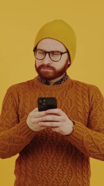 Kafkasyalı hippi gözlüklü ve bereli akıllı telefonunu kullanıyor ve sarı arka plana bakıyor. Akıllı telefon bağımlılığı konsepti. Dikey Çekim.