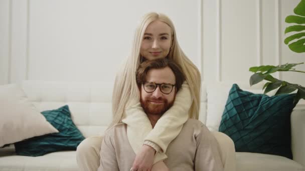 Fiatal gyönyörű európai hosszú hajú szőke nő ölelgeti jóképű férjét hátulról, a kamerába néz — Stock videók