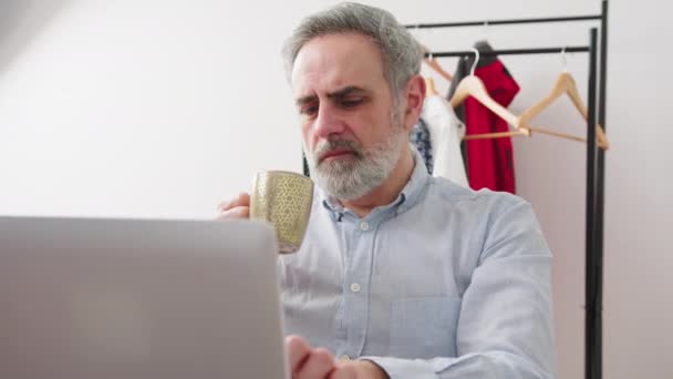 Gray haired bearded male enterprenuer sipping on the coffee while working remotely from home on the laptop — Stock Video