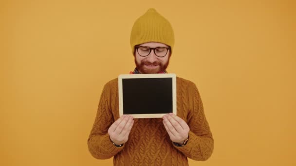 Sorrindo homem hipster barbudo milenar em um gorro mostrando tablet digital com tela preta no estúdio da câmera filmado fundo amarelo — Vídeo de Stock