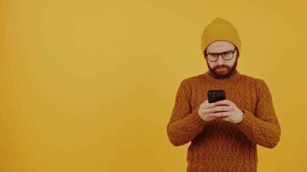 身穿便帽和橙色毛衣的欧洲嬉皮士沉迷于使用他的黑色智能手机，工作室拍摄黄色背景. — 图库视频影像