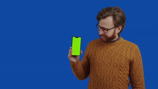 Bonito europeu cara com facial cabelo e óculos olhando e apontando para seu smartphone com verde mock-up tela — Vídeo de Stock