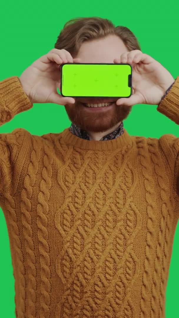 Hombre caucásico barbudo tiro vertical que cubre sus ojos con un teléfono inteligente de pantalla verde sobre fondo verde — Vídeos de Stock