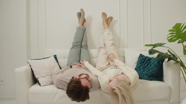 Glückliches Paar in den Zwanzigern, das auf weißer Couch in ihrer nagelneuen Wohnung liegt und die Beine hochlegt und scherzt — Stockvideo