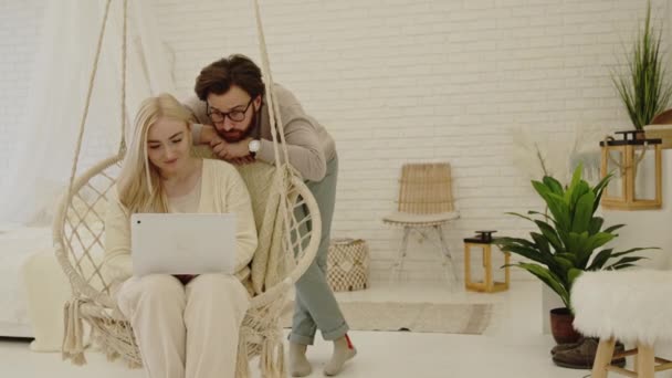Młoda para małżeńska, piękna blondynka millenijna kobieta pokazuje mężowi coś na laptopie — Wideo stockowe