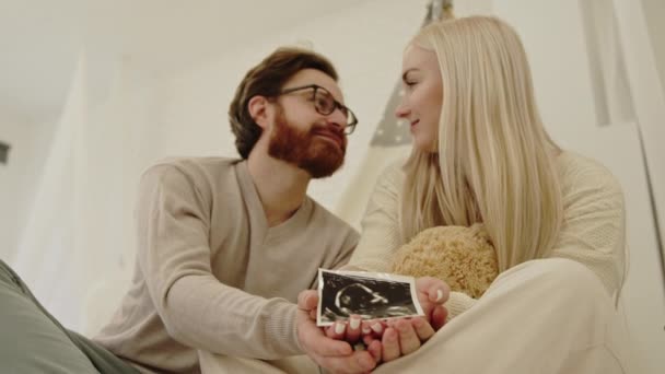 Hamarosan kaukázusi szülők ülnek a padlón, és megmutatják a kamerának a terhesség ultrahangos szkennelését. — Stock videók