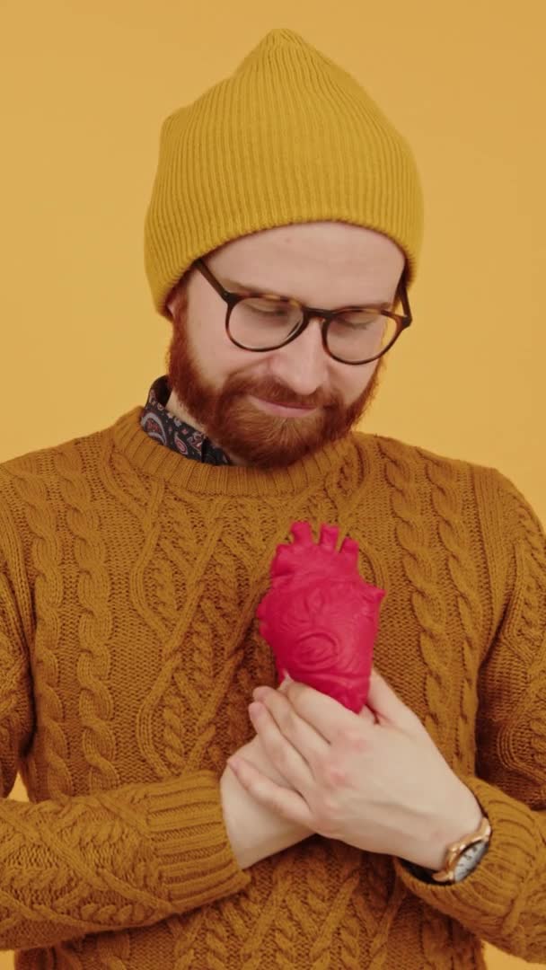 Νεαρή χιλιετή συμπίεση κόκκινο μοντέλο τεχνητή καρδιά με δύο παλάμες, αυτο-φροντίδα και υγιή έννοια της ζωής, κάθετη στούντιο πυροβόλησε κίτρινο φόντο — Αρχείο Βίντεο