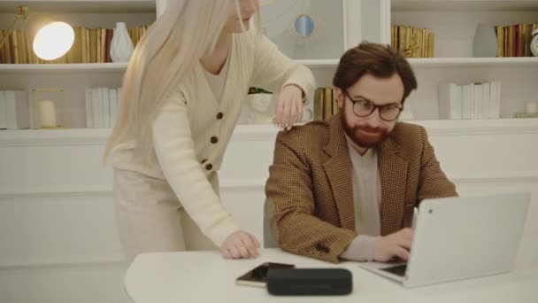Bela mulher caucasiana apoiar seu marido bonito que está sentado na fonte de um laptop branco durante o escritório em casa — Vídeo de Stock