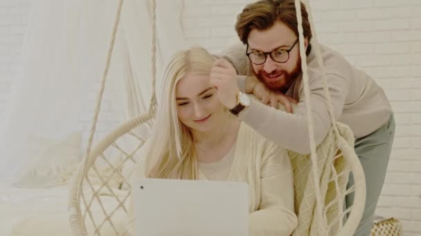 Giovane coppia millenaria guardare video insieme su un computer portatile bianco mentre seduto su un altalena moderna — Video Stock