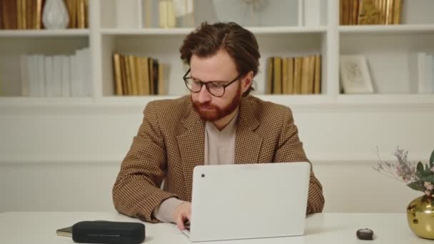 Bonito jovem empresário caucasiano sentado em seu escritório em casa com um laptop, e falando via smartphone. — Vídeo de Stock