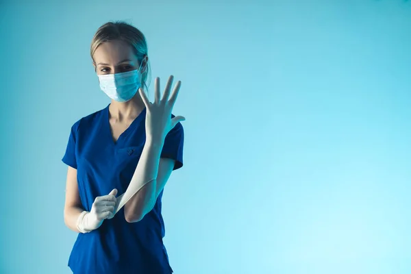 Jovem médica caucasiana vestindo uma luva branca estéril, se preparando para a cirurgia. Médio estúdio tiro, fundo azul. — Fotografia de Stock