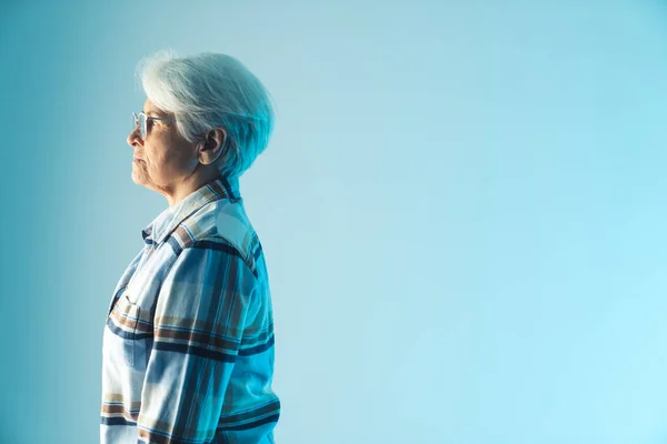 Грустная и задумчивая пожилая серая женщина, стоящая в профиле. Студия Medium снята на голубом фоне. — стоковое фото