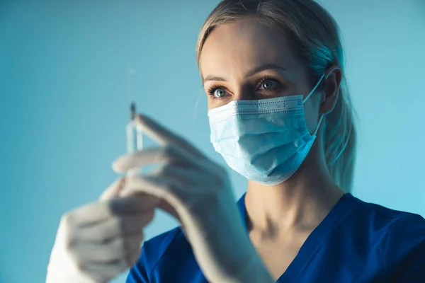 Maskierte kaukasische Praktikantin in der Klinik bereitet Injektion für die Patientin vor. Mittlere Nahaufnahme, blauer Hintergrund. — Stockfoto