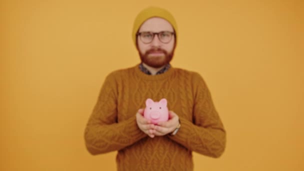 Evropan hipster ukazující jeho růžové prasátko banka na kameru střední studio záběr žluté pozadí — Stock video