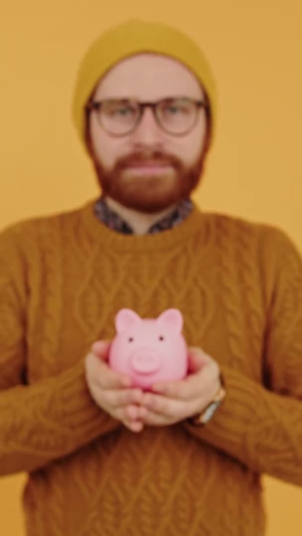 Caucasian hipster man investor showing his piggy bank to camera vertical studio shot yellow background — Stock Video