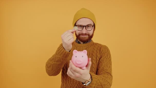 Usmívající se tisíciletý kavkazský hipster muž dává peníze do své prasečí banky, žluté pozadí studia — Stock video