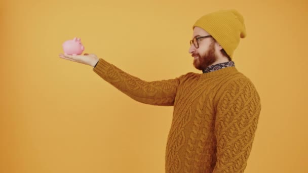 Hipster caucasiano barbudo homem olhando para seu mealheiro e apontando para ele médio estúdio tiro fundo amarelo — Vídeo de Stock