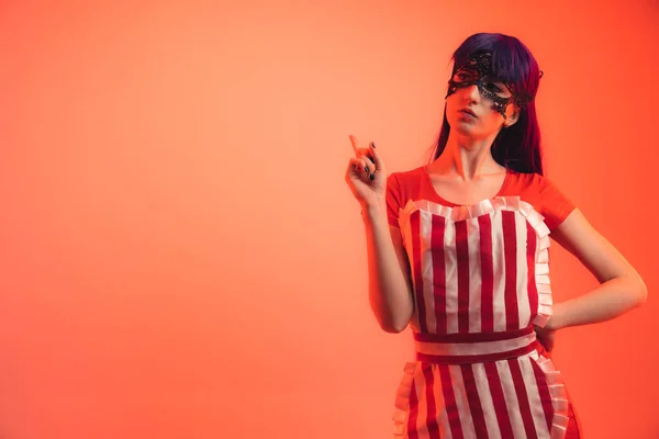 Cosplayer woman in a striped apron and lace mask pointing with her finger at copy space - red background — Stock Photo, Image