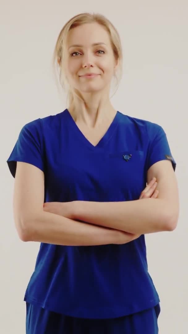 垂直ショット-正のブロンドの医療従事者の女性は、カメラを見て、彼女の腕を横断暗い青の制服で。分離された中間スタジオショット. — ストック動画