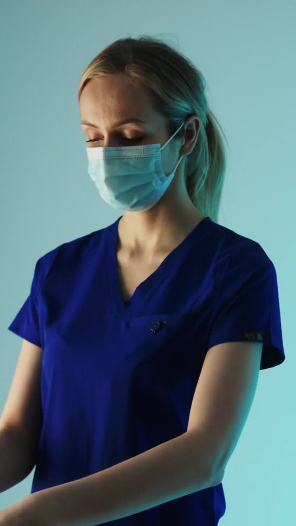 Vídeo Vertical Joven doctora caucásica poniéndose un guante blanco estéril, preparándose para la cirugía. Tiro de estudio medio, fondo azul. — Vídeo de stock