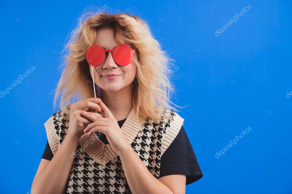 Concetto cabina fotografica. Giovane allegra donna caucasica sorridente  sulla ventina che usa occhiali rossi finti per coprirsi gli occhi. Sfondo  blu studio girato. - Foto Stock: Foto, Immagini © PoppyPix 556837060