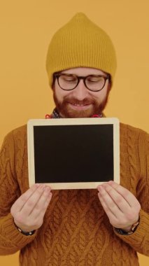  Gözlüklü ve şapkalı genç beyaz adam küçük bir karatahta gösteriyor, ona bakıyor, gülümsüyor ve izole edilmiş fotoğraf stüdyosuna bakıyor sarı arka plan dikey video.