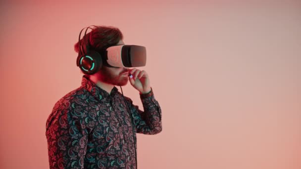 Joven hombre caucásico utiliza gafas de realidad virtual y auriculares, habla en el micrófono y señala su dedo lejos de la pantalla virtual en frente de, vr realidad concepto copia espacio rosa gradiente fondo — Vídeo de stock