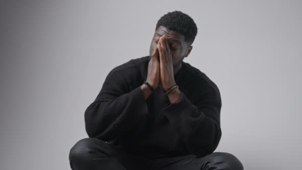 Ung svart man ser bekymrad sätta huvudet i sina händer som ett tecken på oro, grå bakgrund — Stockvideo