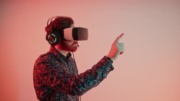 Joven hombre caucásico atractivo en gafas de realidad virtual y auriculares, pasa con el dedo en la pantalla, vr realidad concepto copia espacio rosa gradiente fondo aislado — Vídeo de stock