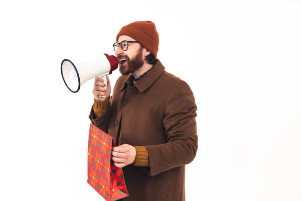 Retrato de un joven guapo hombre caucásico gritando en el megáfono y sosteniendo la bolsa de Navidad roja vestimenta casual tonos de ropa marrón fondo blanco aislado copia espacio estudio disparo —  Fotos de Stock