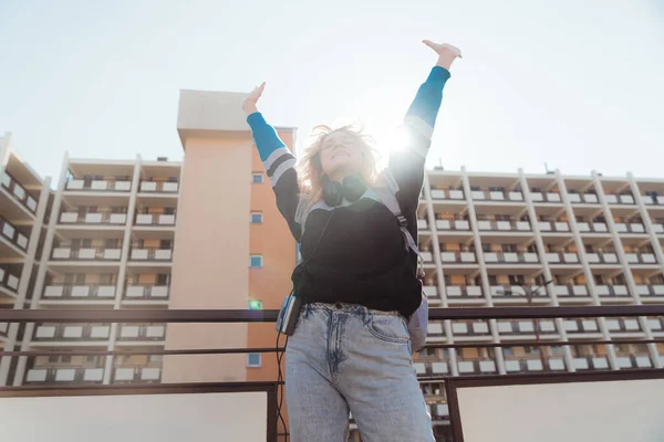 Den sorglösa 90-talsinspirerade kaukasiska flickan kastar händerna mot den blå himlen. Block av lägenheter i bakgrunden. — Stockfoto