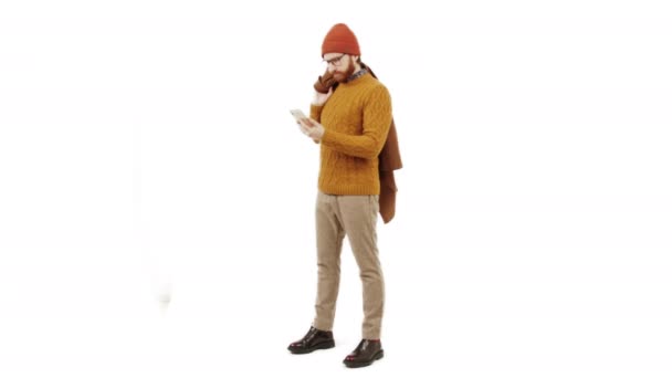 Молодой красивый бородатый белый мужчина смотрит на свой смартфон и улыбается, держа пальто на плече повседневной одежде белый фоновый студия выстрелил полный снимок пространство для копирования — стоковое видео