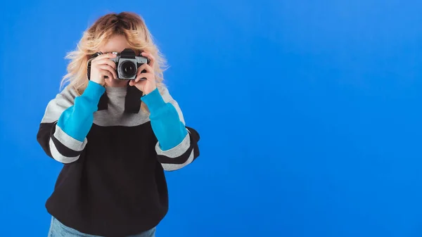 中等摄影棚拍摄的金发90年代风格的女孩在蓝色背景上用老式模拟相机拍照. — 图库照片