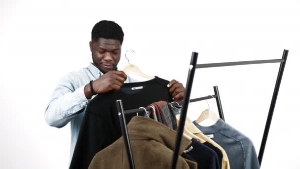 Video schöner afroamerikanischer Mann wählt einen Pullover von der Kleiderstange horizontal medium shot weißen Hintergrund — Stockvideo