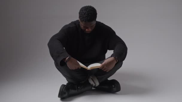 Video Afro-Amerikaanse man het lezen van een boek op de vloer in studio veranderende licht verlichting volledige shot geïsoleerde horizontale kopieerruimte grijze achtergrond — Stockvideo