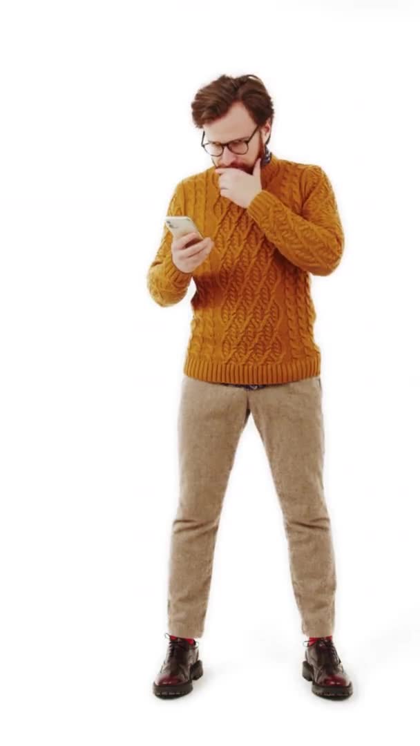 Вертикальное видео молодой сосредоточенный белый мужчина использует свой телефон и касается бороды белый фон изолированы полный снимок — стоковое видео