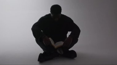 Siyah, yakışıklı bir erkek, yerde oturup kafasını sallarken kitap okuyor. Mutluluğun işareti olarak, gri arka plan.