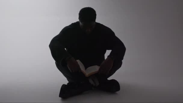 Negro bien parecido macho leer un libro mientras está sentado en el suelo y asintiendo con la cabeza como un signo de felicidad, fondo gris — Vídeo de stock