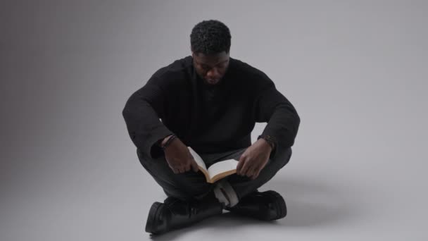 Μαύρος όμορφος άντρας διαβάζει ένα βιβλίο ενώ κάθεται στο έδαφος και μετά κοιτάζει επίμονα την κάμερα, σκούρο φόντο, μεσαίο κοντινό πλάνο. — Αρχείο Βίντεο