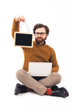 Genç, yakışıklı, beyaz bir adam önünde küçük bir karatahta tutuyor dizüstü bilgisayarını kucağında yerde oturuyor, bacak bacak üstüne atıyor.