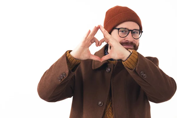 Retrato de joven hombre caucásico barbudo en gafas y sombrero sonriendo y mostrando gesto de corazón con sus manos hipster marrón ropa casual aislado fondo blanco estudio disparo — Foto de Stock