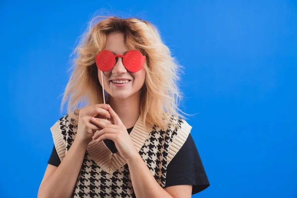 Playful European young woman grinning and covering her eyes with fake red glasses over blue background. Isolated studio shot. Party concept. — Stock Photo, Image