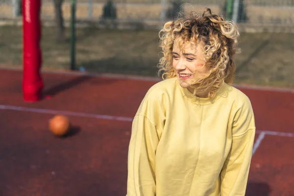 Attraktiv nöjd blond europeisk kvinna står på basketplanen, tittar bort och ler, blåsigt väder kopia utrymme medium shot — Stockfoto