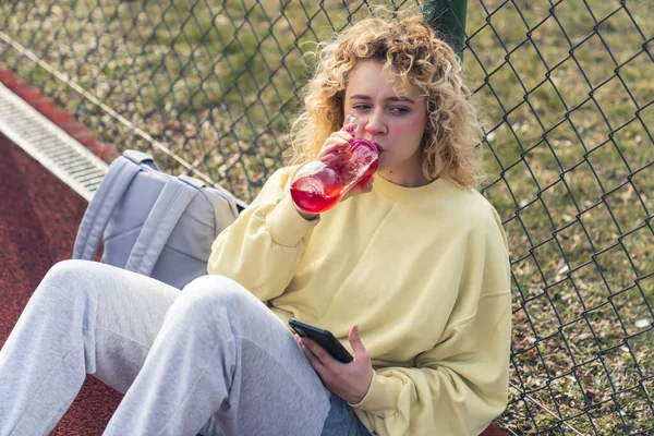 Maçtan sonra su içen genç sarışın kadın, spor sahasında oturuyor, telefonu tutuyor ve orta mesafeden bakıyor. 